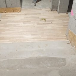 Fragment wnętrza w trakcie prac wykończeniowych: ułożona drewniana podłoga przechodzi w surowy beton schodów. W tle drabina oparta o betonowy filar.