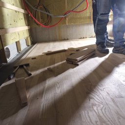 Układanie drewnianej podłogi w pomieszczeniu z ocieplonymi ścianami. Widoczne narzędzia i fragmenty desek. Dwóch pracowników w jeansach w tle.