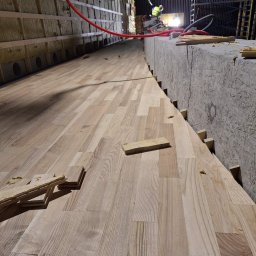 Układanie parkietu w hali: jasne deski, widoczne fragmenty izolacji ścian i pracujący w oddali monter. Ujęcie z niskiej perspektywy podkreśla długość pomieszczenia.