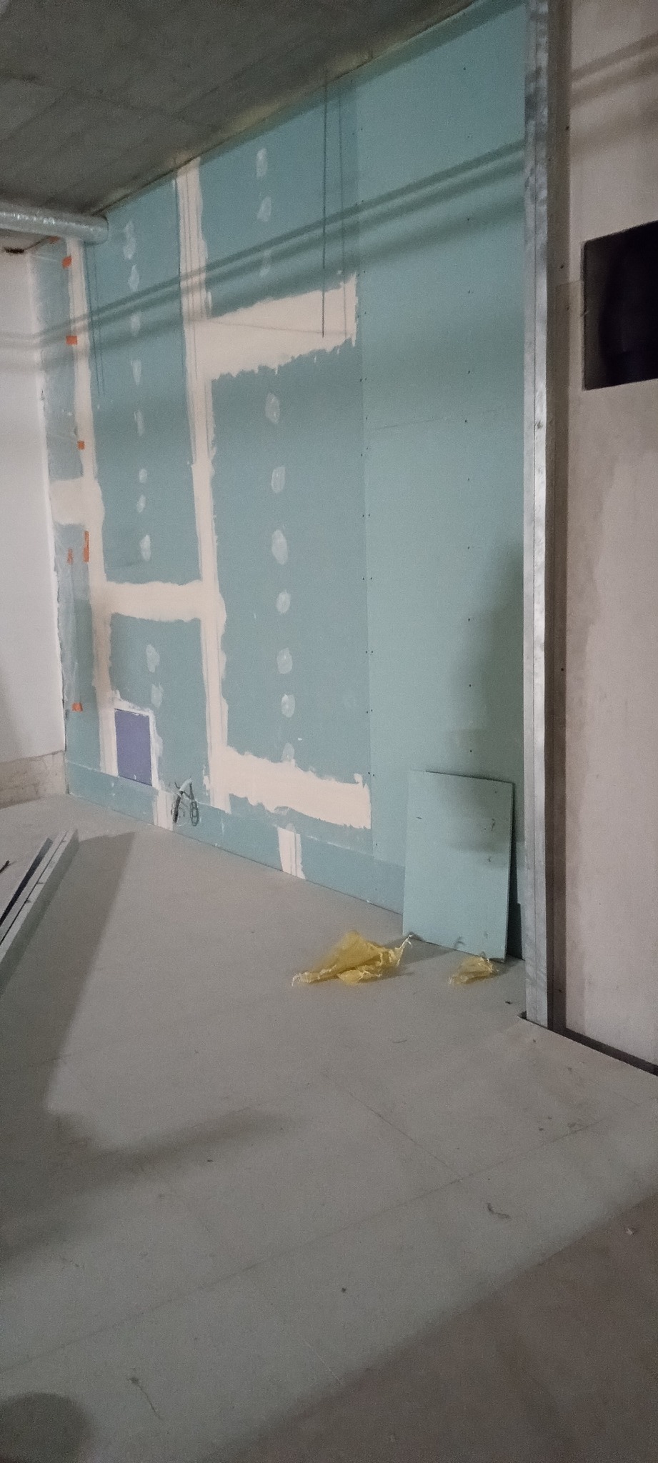 Ściana z zamontowaną płytą gipsowo-kartonową, widoczne profile i miejsca łączeń. Na podłodze leżą resztki folii. Surowy stan wykończenia wnętrza.