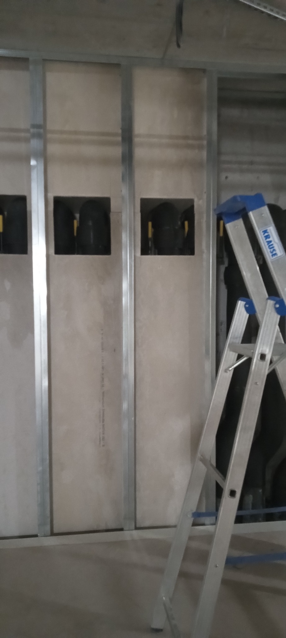 Montaż płyty karton gips na metalowym stelażu z widocznymi otworami na instalacje. W tle drabina Krause. Prace wykończeniowe w trakcie realizacji.