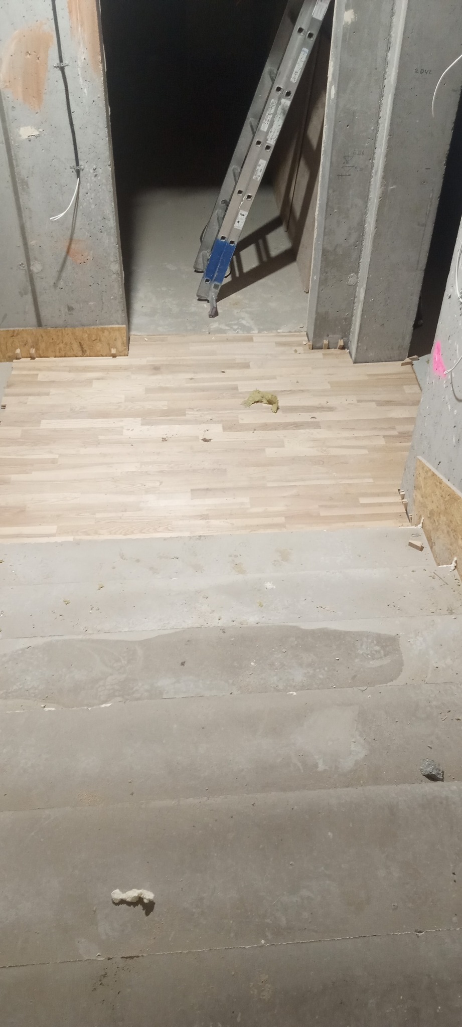 Fragment wnętrza w trakcie prac wykończeniowych: ułożona drewniana podłoga przechodzi w surowy beton schodów. W tle drabina oparta o betonowy filar.