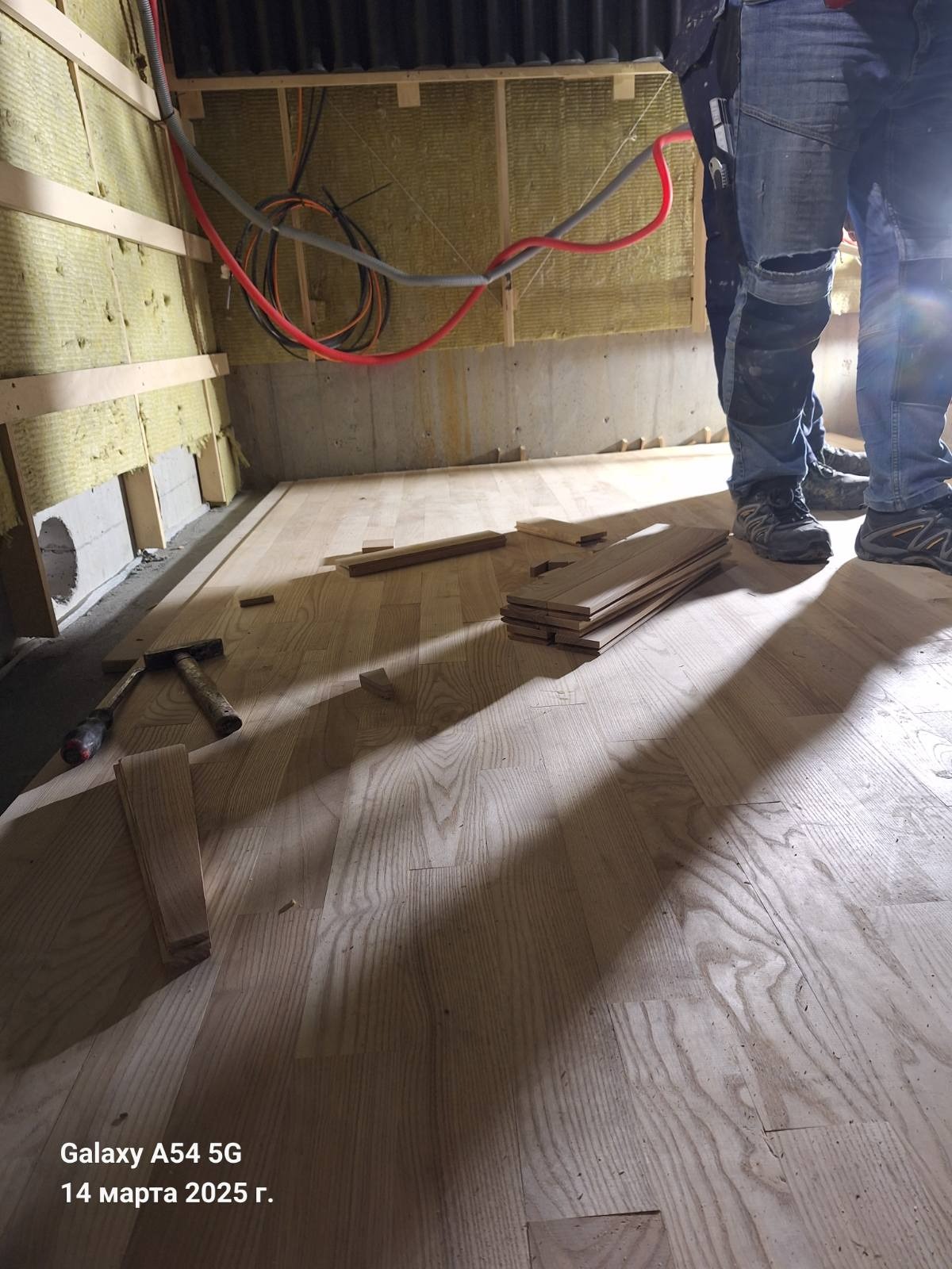 Układanie drewnianej podłogi w pomieszczeniu z ocieplonymi ścianami. Widoczne narzędzia i fragmenty desek. Dwóch pracowników w jeansach w tle.