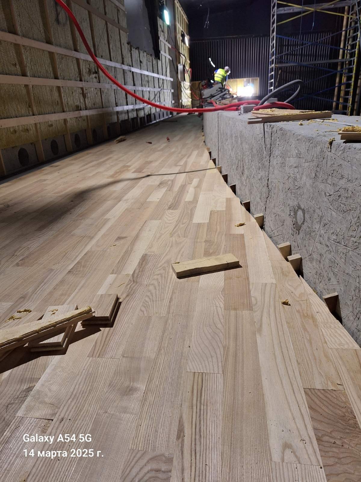 Układanie parkietu w hali: jasne deski, widoczne fragmenty izolacji ścian i pracujący w oddali monter. Ujęcie z niskiej perspektywy podkreśla długość pomieszczenia.