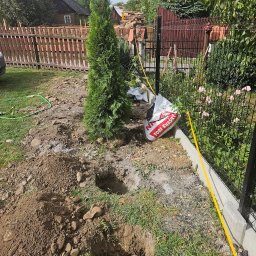 Allgarden Usługi Ogrodnicze Piotr Serwin - Przygotowane dołki i tuje w ogrodzie, torba z torfem obok. Etap sadzenia drzewek iglastych przy ogrodzeniu, w tle drewniany dom i porozrzucane deski.