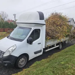 Biały samochód dostawczy z wysoką zabudową, wypełniony po brzegi gałęziami i liśćmi, stojący na żwirowej drodze w jesiennej scenerii.