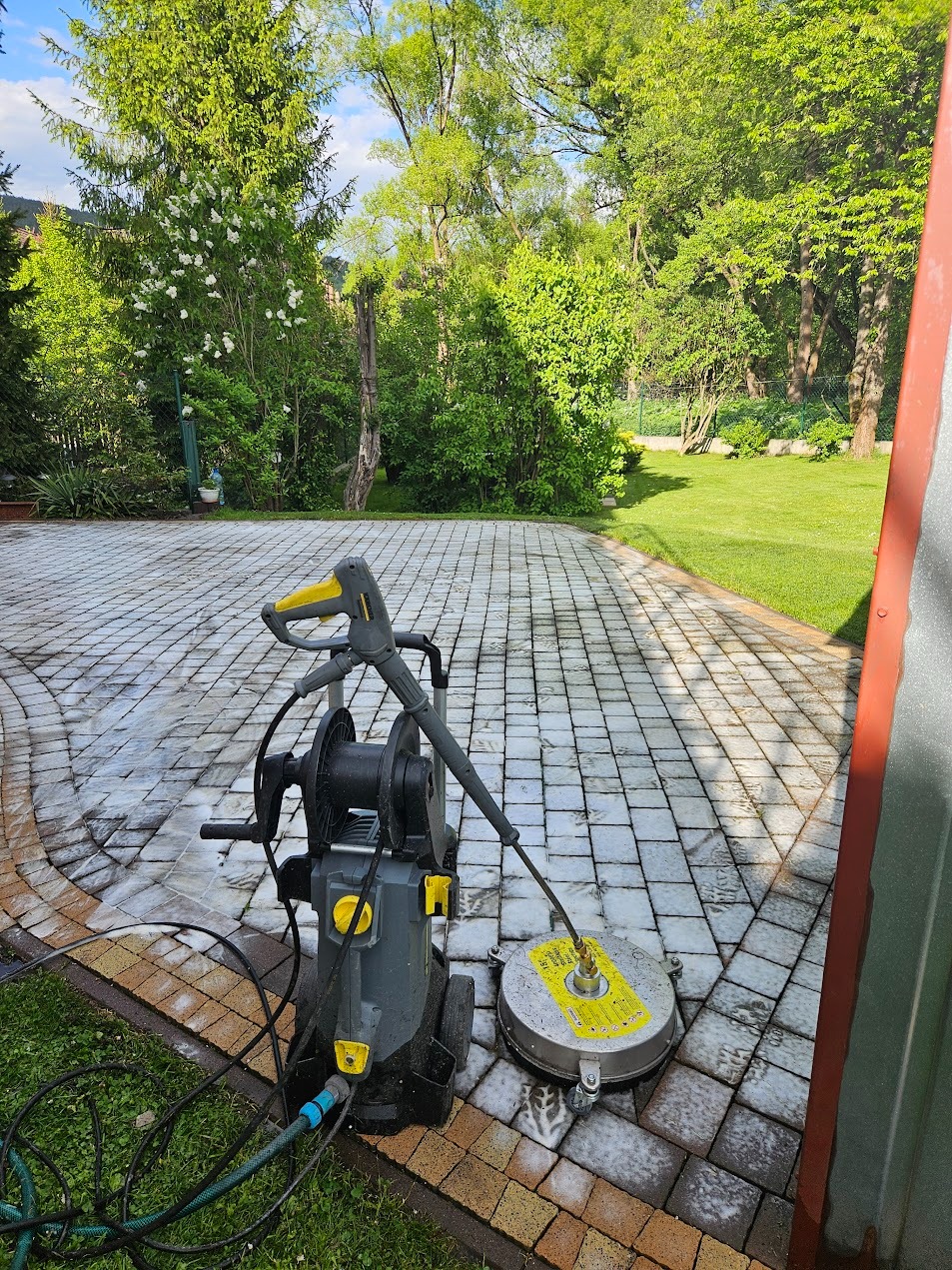 Mycie kostki brukowej myjką ciśnieniową Karcher z okrągłą dyszą, widoczne mokre i czyste fragmenty kostki. Trawa i zieleń w tle. Słoneczny dzień.