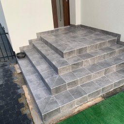 KORTIN - Wykończone płytkami schody wejściowe do domu, z widoczną fugą i fragmentem trawnika. Obok widoczny fragment ogrodzenia i kostki brukowej.