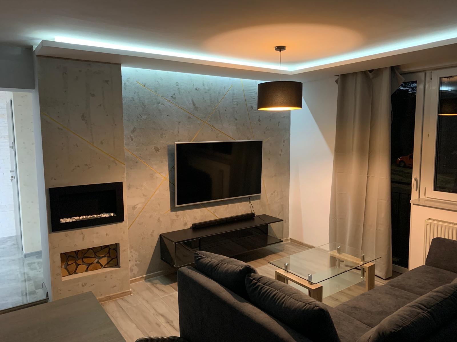 Salon po remoncie: ściana z efektem betonu, dekoracyjne złote linie, kominek elektryczny z drewnem, wiszący telewizor, szara sofa i szklany stolik kawowy.