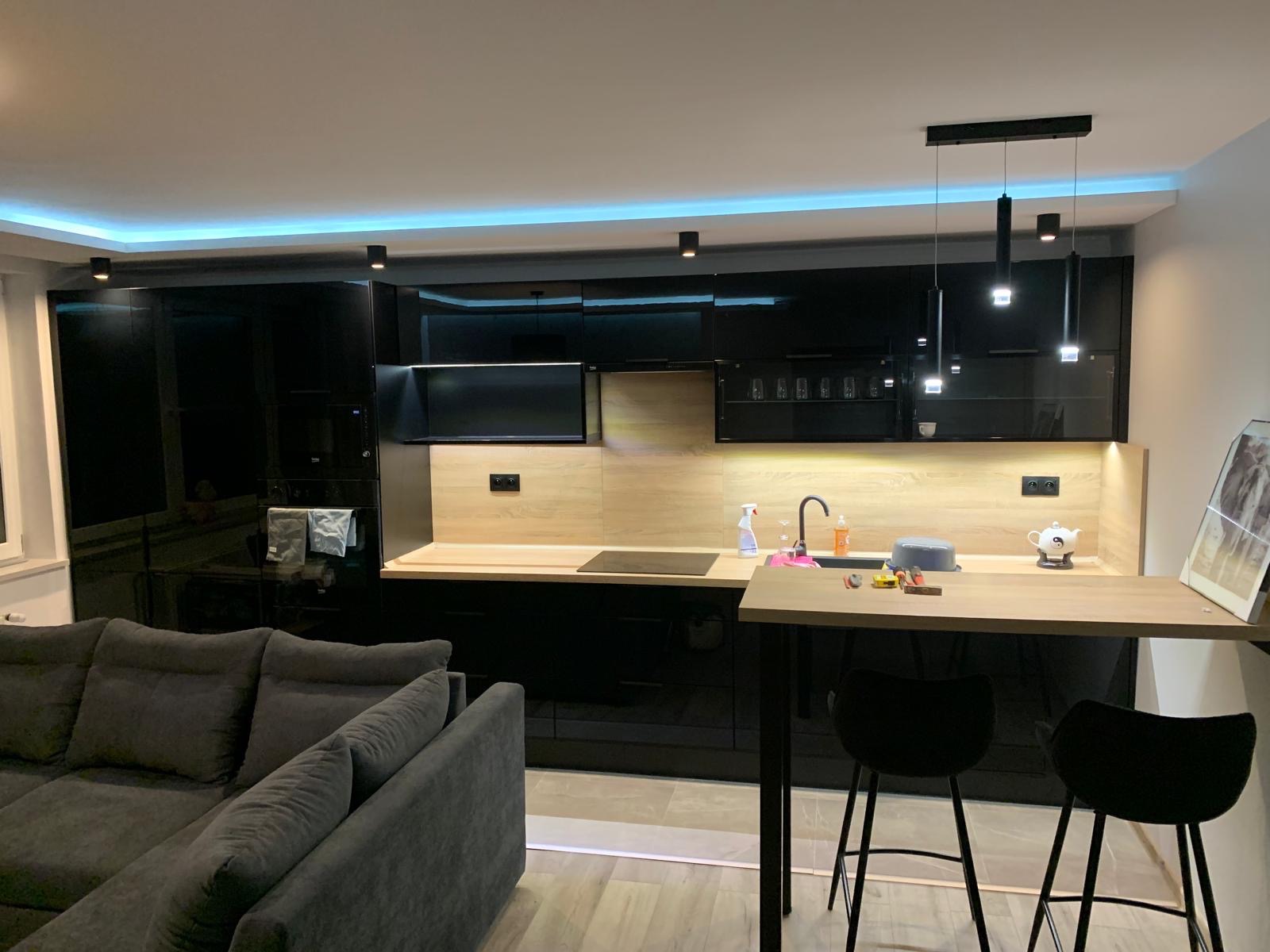 Nowoczesna kuchnia z czarnymi, błyszczącymi szafkami i drewnianym blatem, oświetlona ledami. Widoczny fragment salonu z szarą sofą i barkiem z krzesłami.