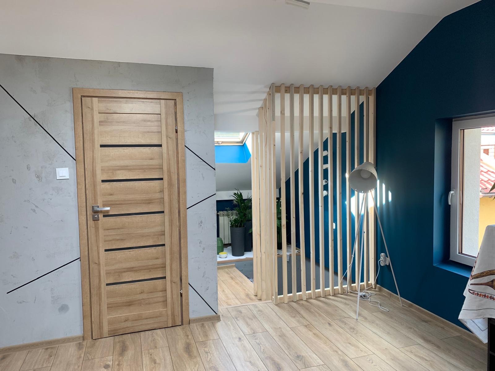 Drewniane drzwi z poziomymi, czarnymi paskami na tle szarej ściany z czarnymi liniami. Widoczny ażurowy przepierzenie i fragment granatowej ściany z oknem.