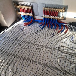 WaterTamer Michał Bełdycki - Rozłożone rury ogrzewania podłogowego na izolacji, z podłączonymi rozdzielaczami z rurami czerwonymi i niebieskimi. Widok z góry na instalację w surowym wnętrzu.