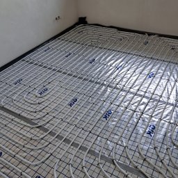 WaterTamer Michał Bełdycki - Białe rury ogrzewania podłogowego ułożone na folii w Ostrołęce, widoczne przyłącza na ścianie. Instalacja w trakcie realizacji, bez wylewki.