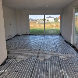 WaterTamer Michał Bełdycki - Białe rury ogrzewania podłogowego ułożone na folii w jasnym pomieszczeniu z dużym oknem przesuwnym i widokiem na domy w tle.