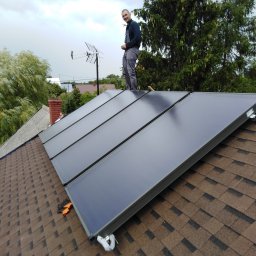 WaterTamer Michał Bełdycki - Instalacja solarna na dachu z brązowej dachówki bitumicznej. Instalator stoi na panelach, uśmiecha się. W tle drzewa i antena telewizyjna.