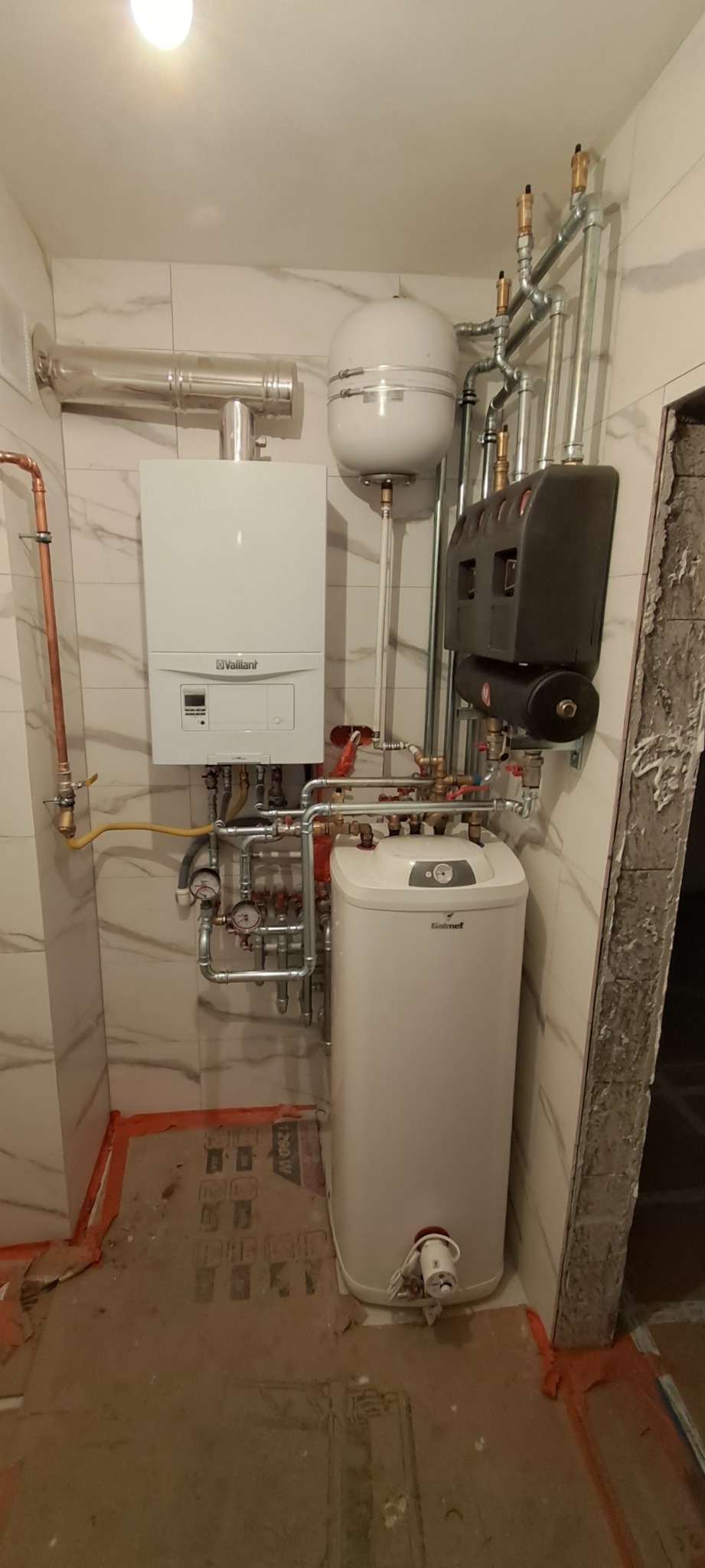 Zestaw kotła gazowego Vaillant z zasobnikiem Galmet i rozbudowaną instalacją hydrauliczną w pomieszczeniu wykończonym marmurową płytką. Widok z dołu.