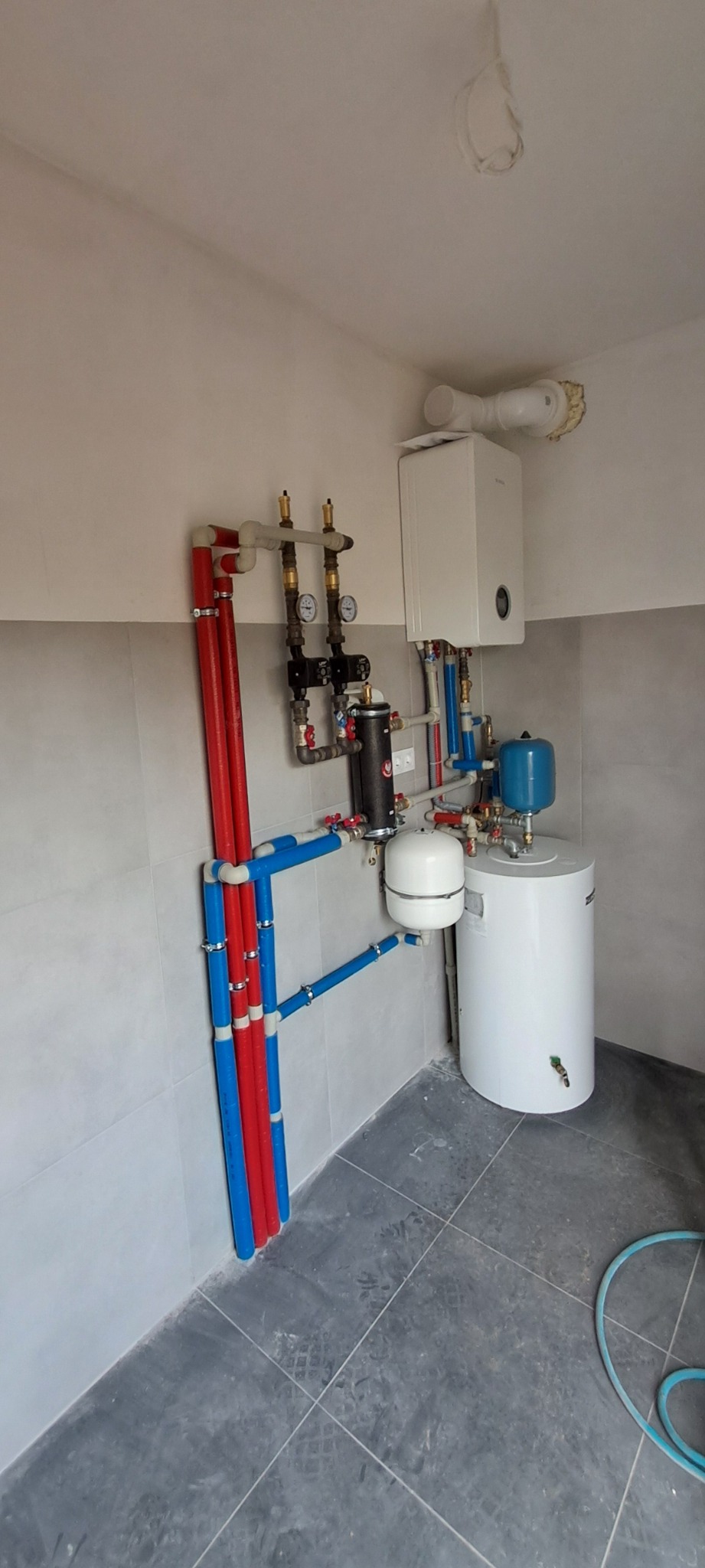 Kompletna instalacja grzewcza z piecem gazowym, zasobnikiem, naczyniami przeponowymi i rurami z tworzyw sztucznych w kotłowni na szarych płytkach.