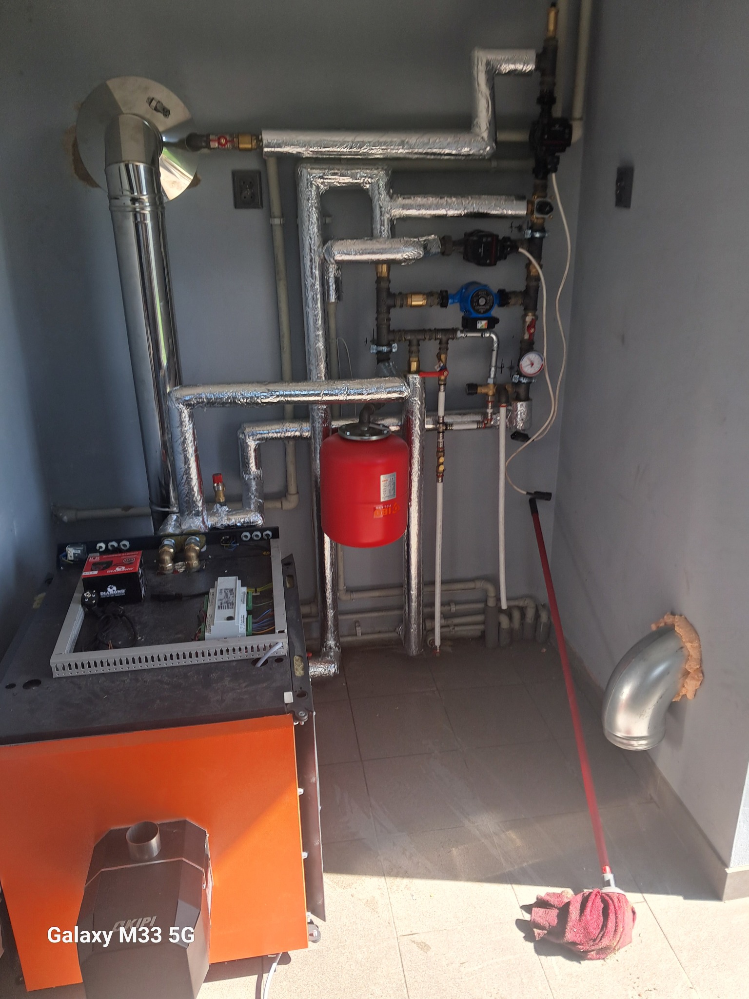 Pomarańczowy piec z podłączonymi rurami, zaworami i osprzętem w szarej kotłowni. Widoczny mop na podłodze i fragment ściany z otworem.
