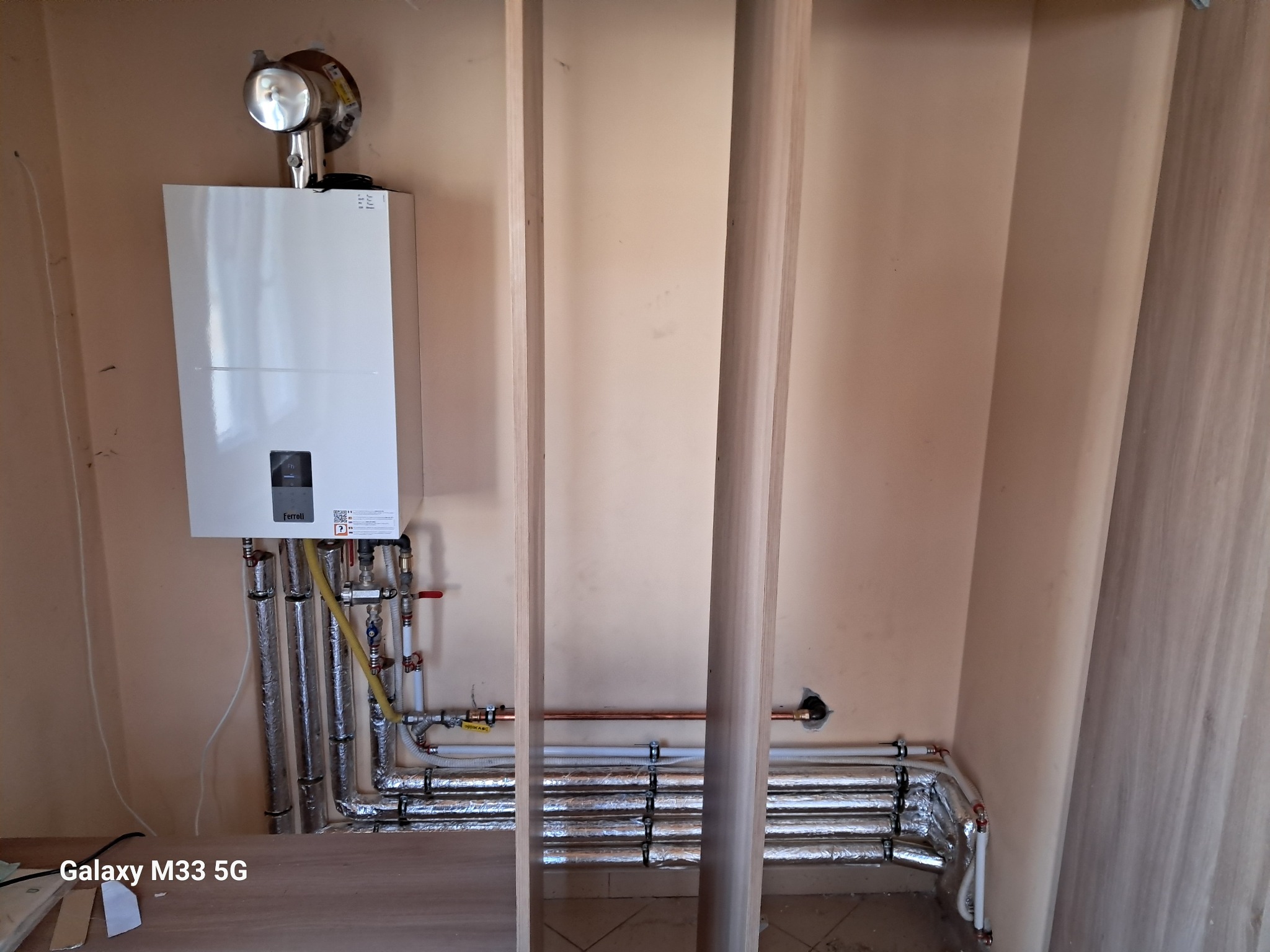 Biały piec gazowy Ferolli z podłączeniami na ścianie o beżowym odcieniu. Widoczne rury w izolacji i elementy instalacji. Ujęcie z perspektywy podłogi.