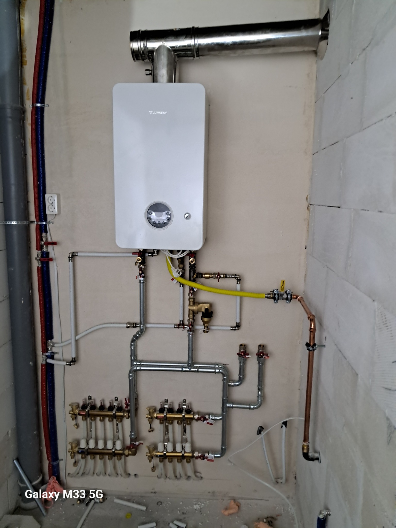Biały piec gazowy Junkers z podłączeniami: rury miedziane, stalowe i plastikowe na ścianie z betonu komórkowego. Widoczny rozdzielacz ogrzewania podłogowego.