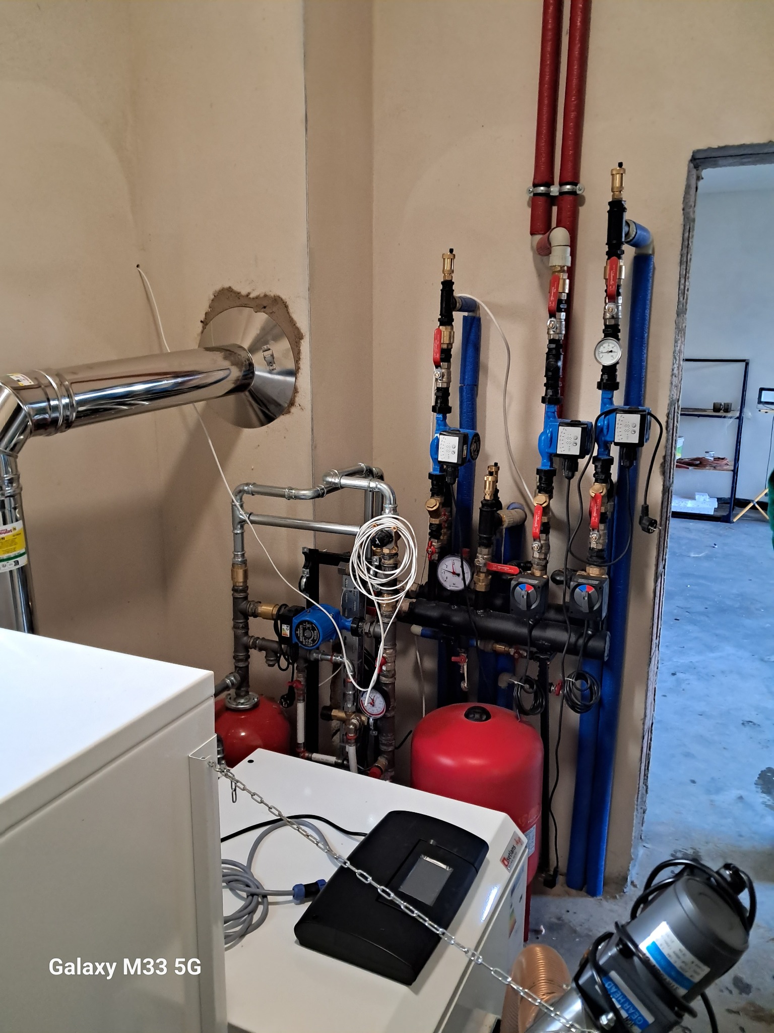 Węzeł cieplny w kotłowni: biały piec, czerwone zbiorniki, niebieskie rury, zawory i manometry. Widoczne elementy instalacji grzewczej w surowym pomieszczeniu.