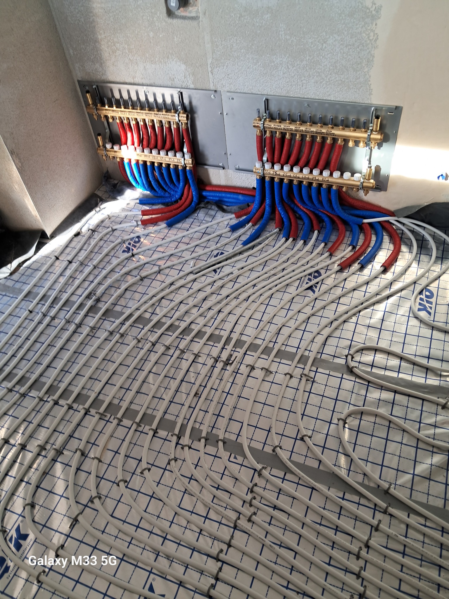 Rozłożone rury ogrzewania podłogowego na izolacji, z podłączonymi rozdzielaczami z rurami czerwonymi i niebieskimi. Widok z góry na instalację w surowym wnętrzu.