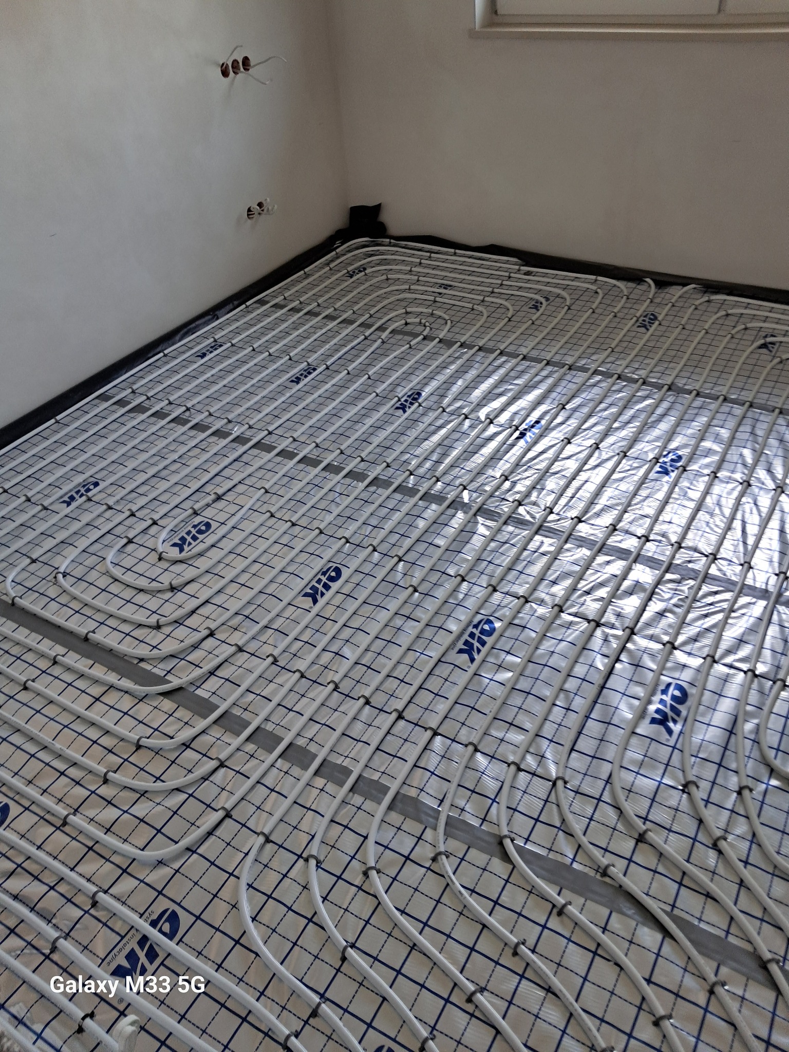 Białe rury ogrzewania podłogowego ułożone na folii w Ostrołęce, widoczne przyłącza na ścianie. Instalacja w trakcie realizacji, bez wylewki.