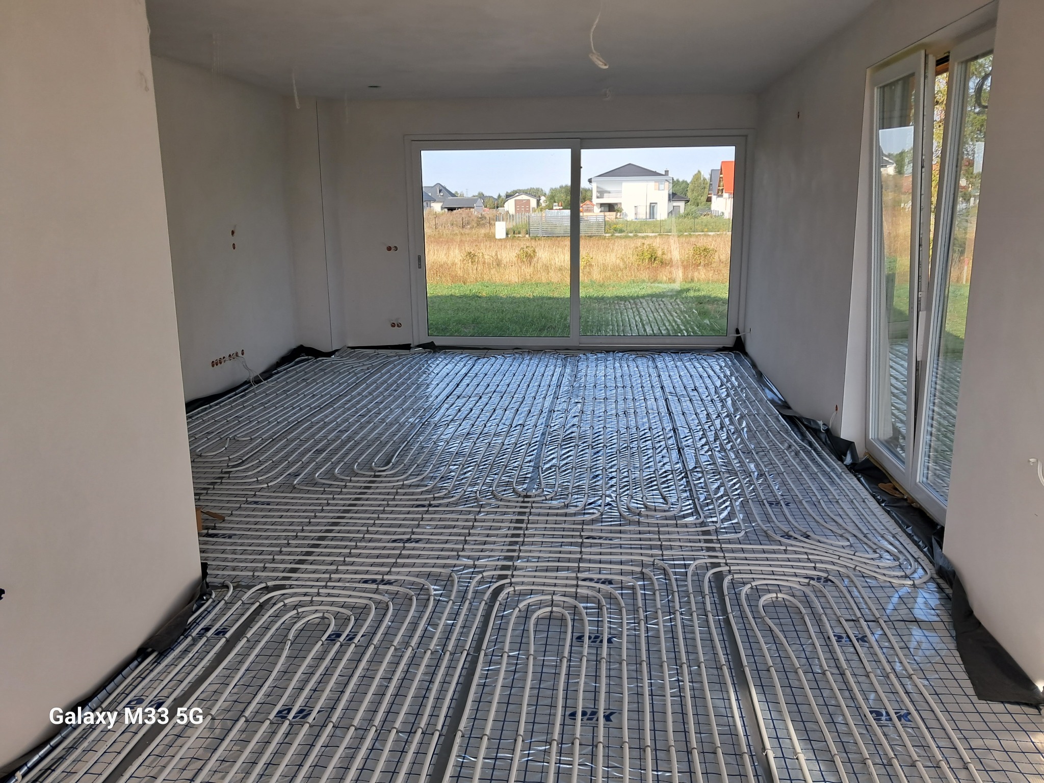 Białe rury ogrzewania podłogowego ułożone na folii w jasnym pomieszczeniu z dużym oknem przesuwnym i widokiem na domy w tle.