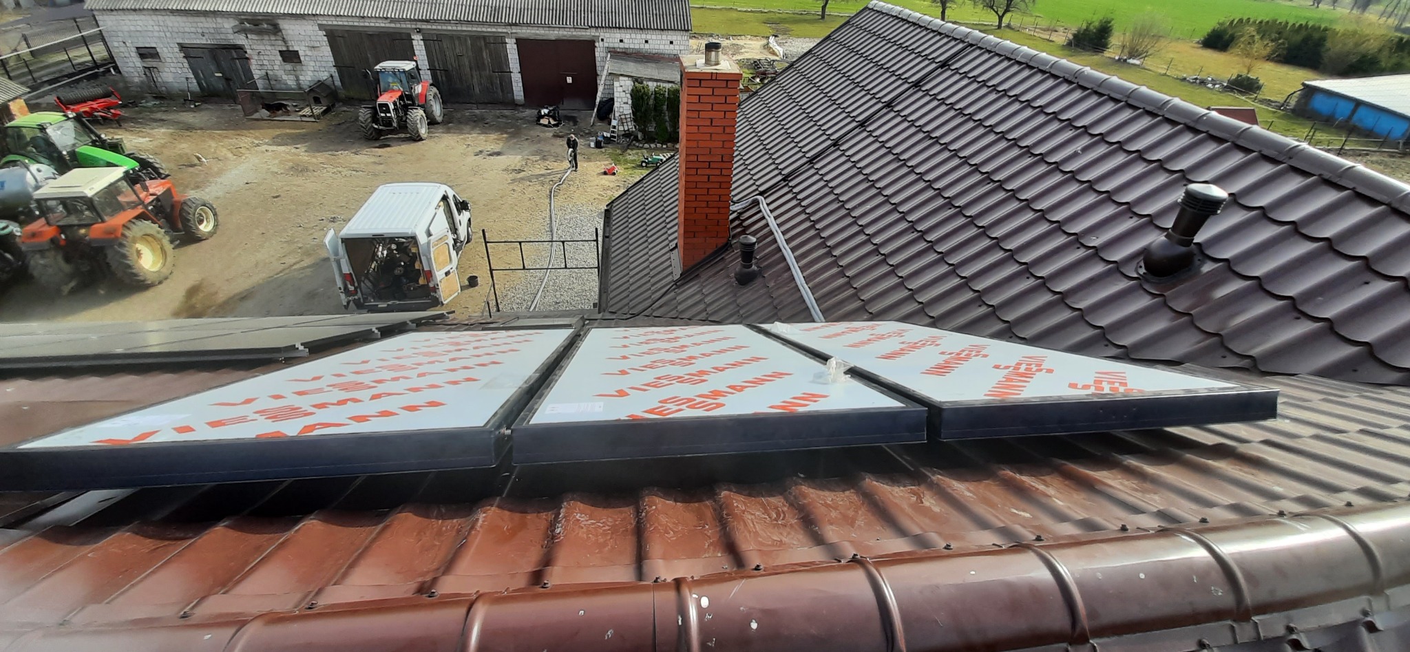 Panele słoneczne Viessmann na brązowym dachu, widok z góry. W tle budynki gospodarcze, ciągniki i biały bus. Ujęcie z perspektywy dachu.