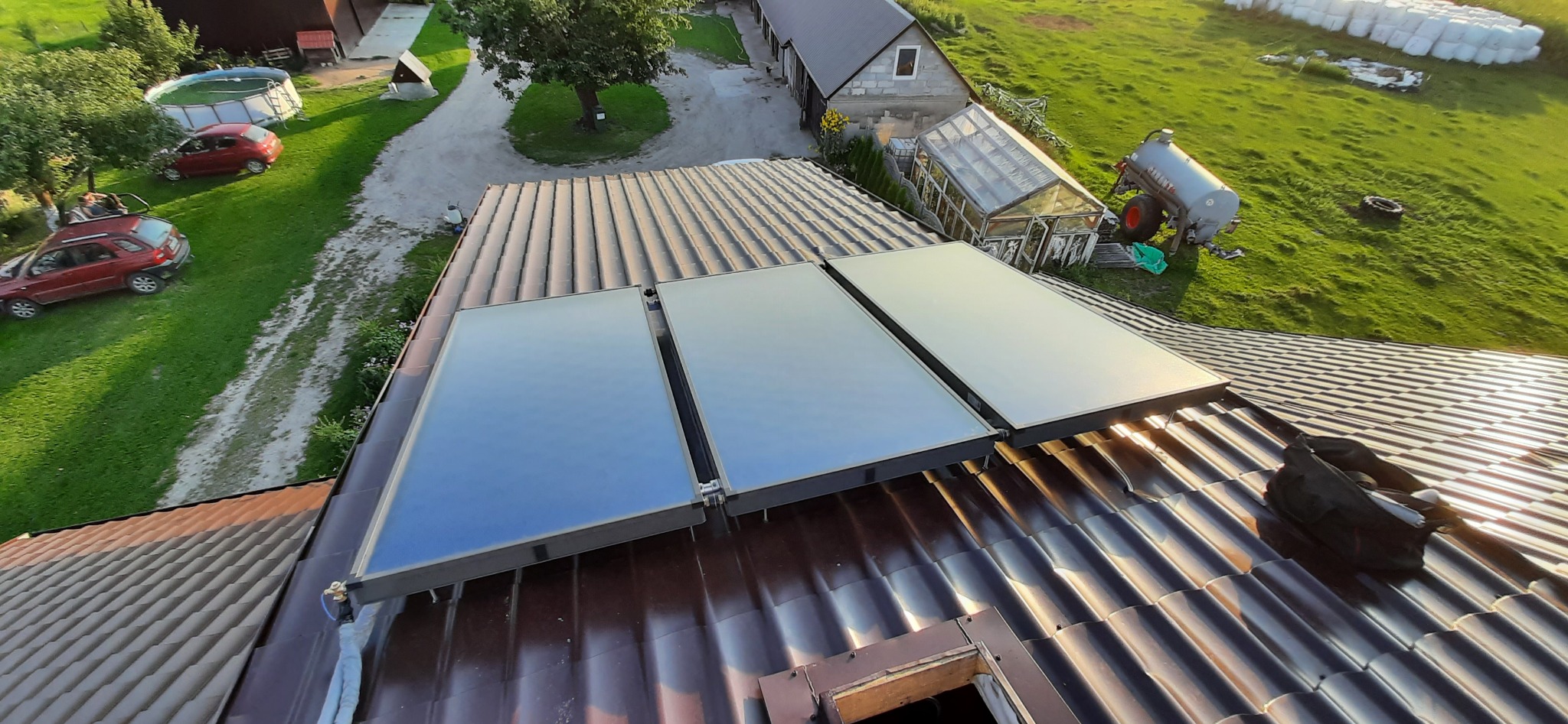 Trzy panele solarne zamontowane na brązowym dachu z blachodachówki, widok z góry na wiejski krajobraz z budynkami gospodarczymi i zielonym terenem.