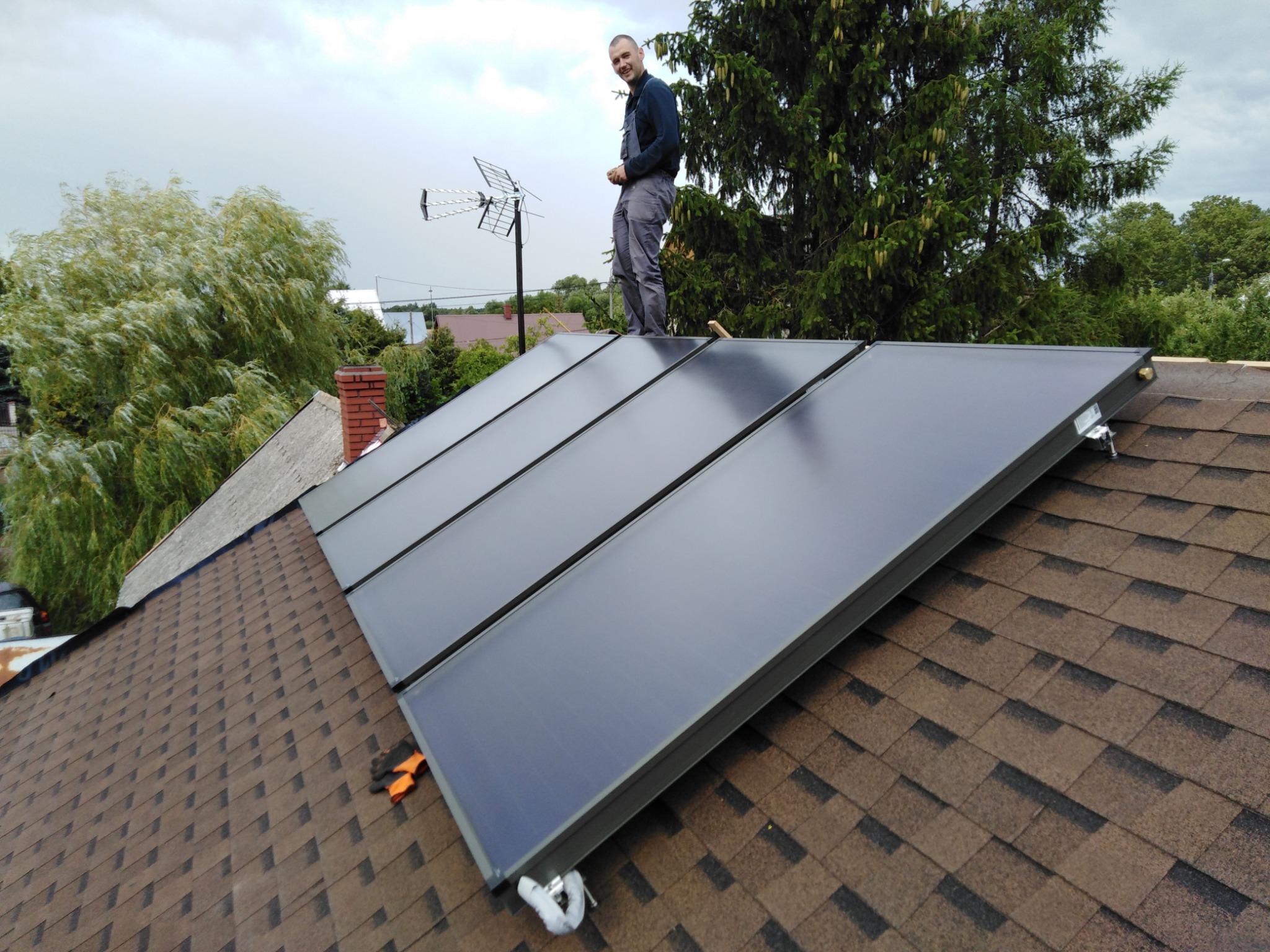 Instalacja solarna na dachu z brązowej dachówki bitumicznej. Instalator stoi na panelach, uśmiecha się. W tle drzewa i antena telewizyjna.