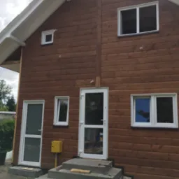 Brunatna elewacja domu z bali, białe ramy okien i drzwi, betonowe schody wejściowe w trakcie budowy, widoczny fragment instalacji gazowej i narzędzi.