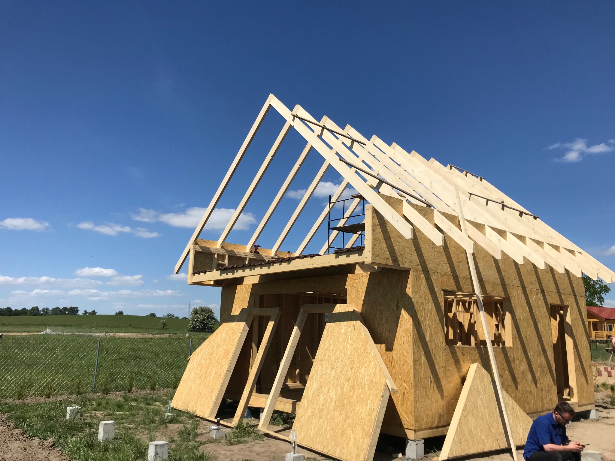Konstrukcja domu szkieletowego w trakcie budowy, widoczny szkielet dachu i ścian z płyt OSB, na tle błękitnego nieba. W pobliżu siedzi osoba.