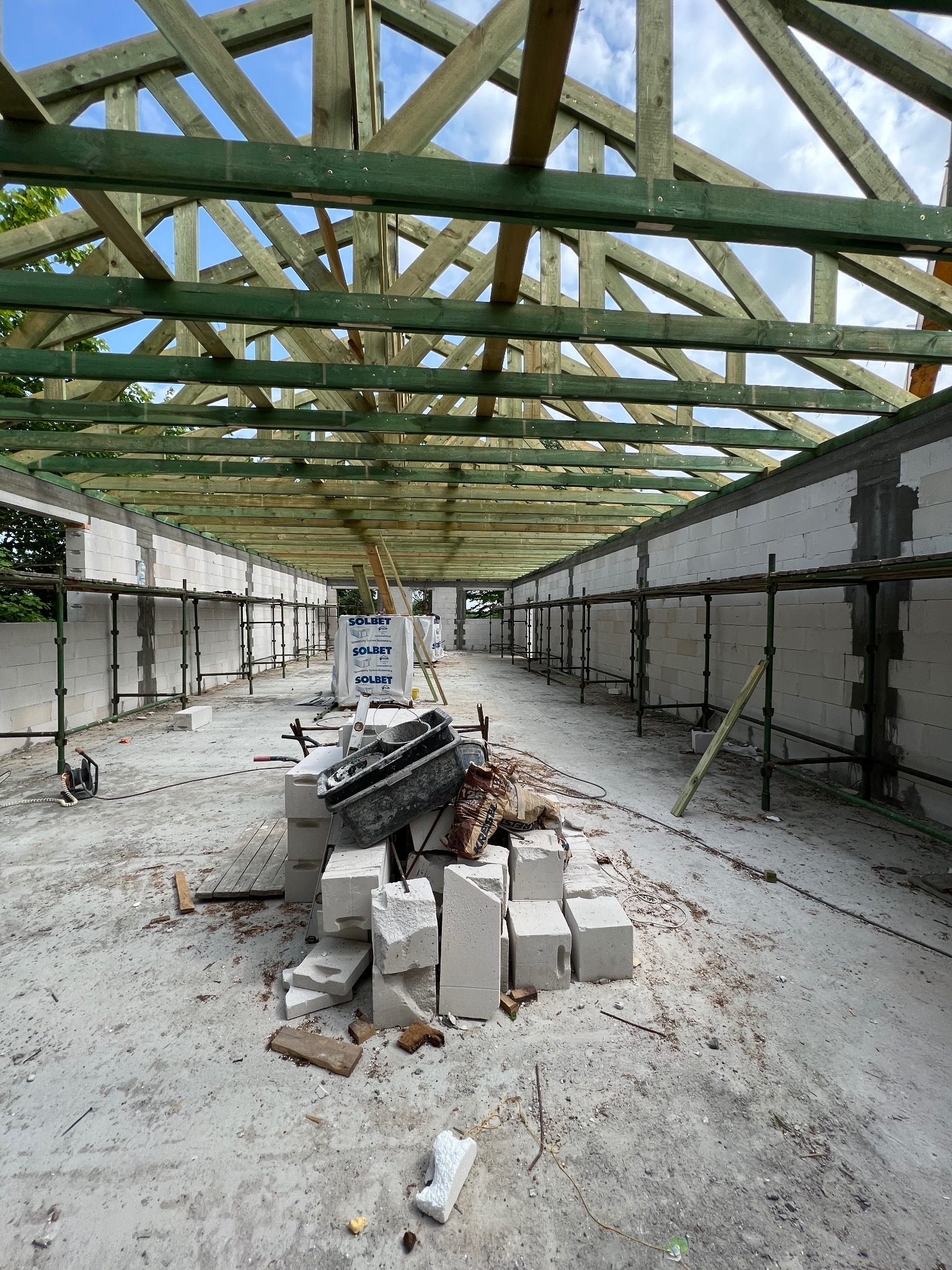 Wnętrze budowy: ściany z betonu komórkowego, zielona drewniana konstrukcja dachu, rusztowania, worki i gruz. Widok na budowę w trakcie realizacji.