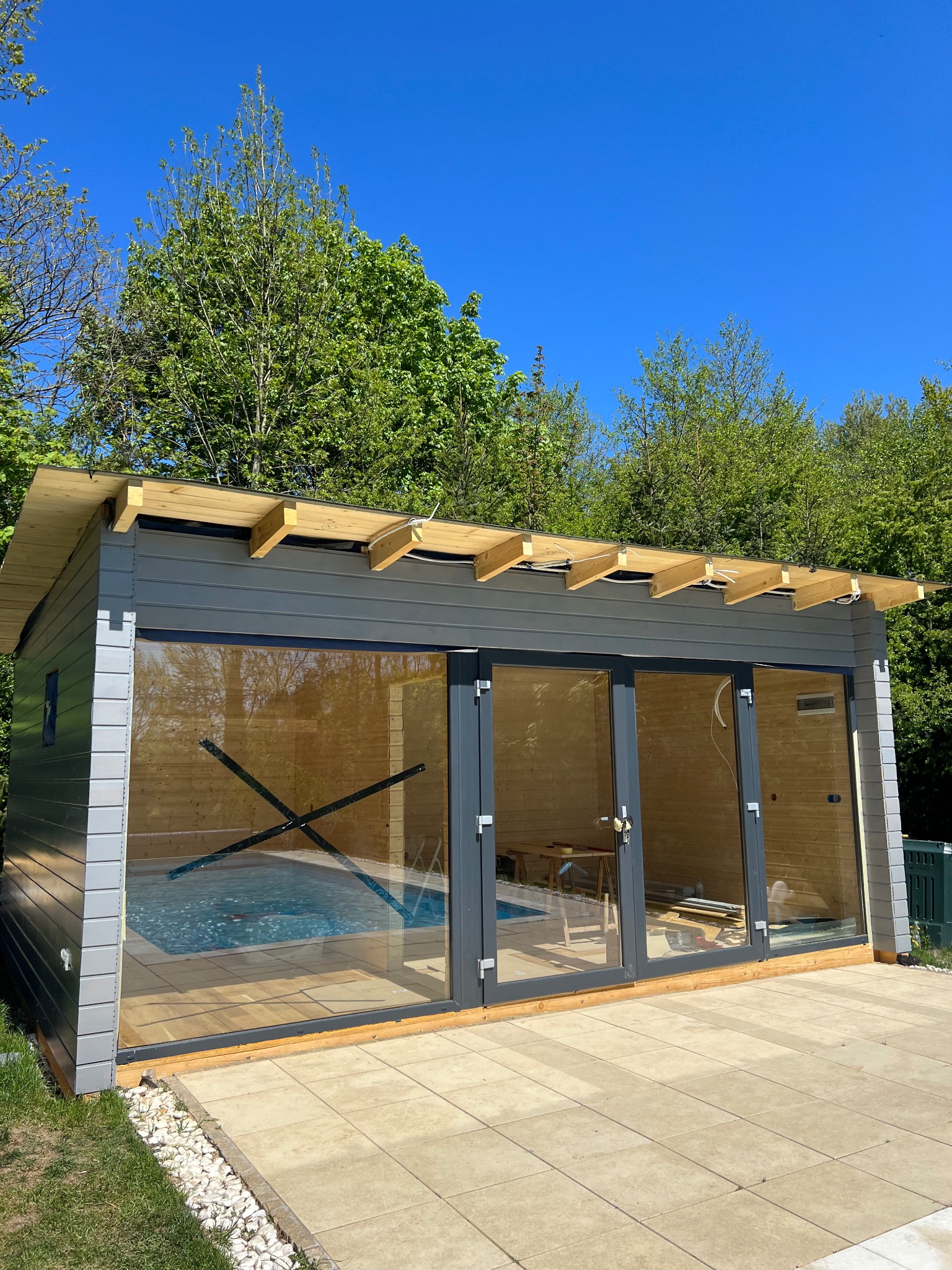 Nowoczesna, szara altana z dużymi przeszkleniami i basenem w środku. Uwagę zwraca drewniana konstrukcja dachu i kamienna ścieżka wokół.