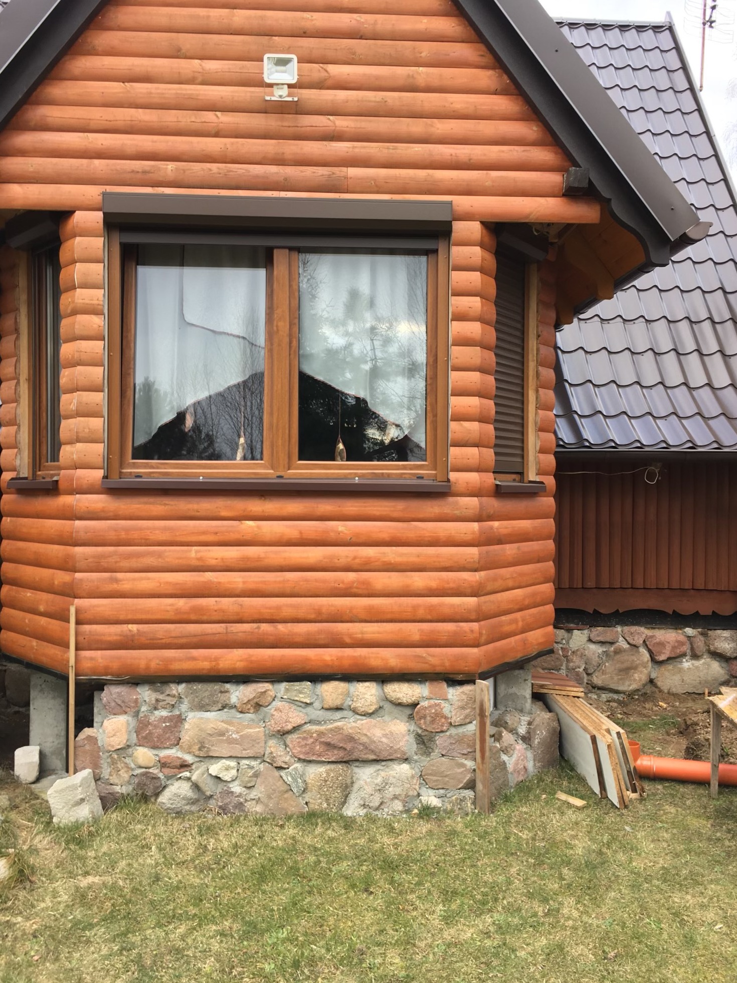 Fragment domu z bali z oknami i kamienną podmurówką w Elblągu. Widoczne elementy wykończeniowe i fragment dachu z brązowej blachodachówki.