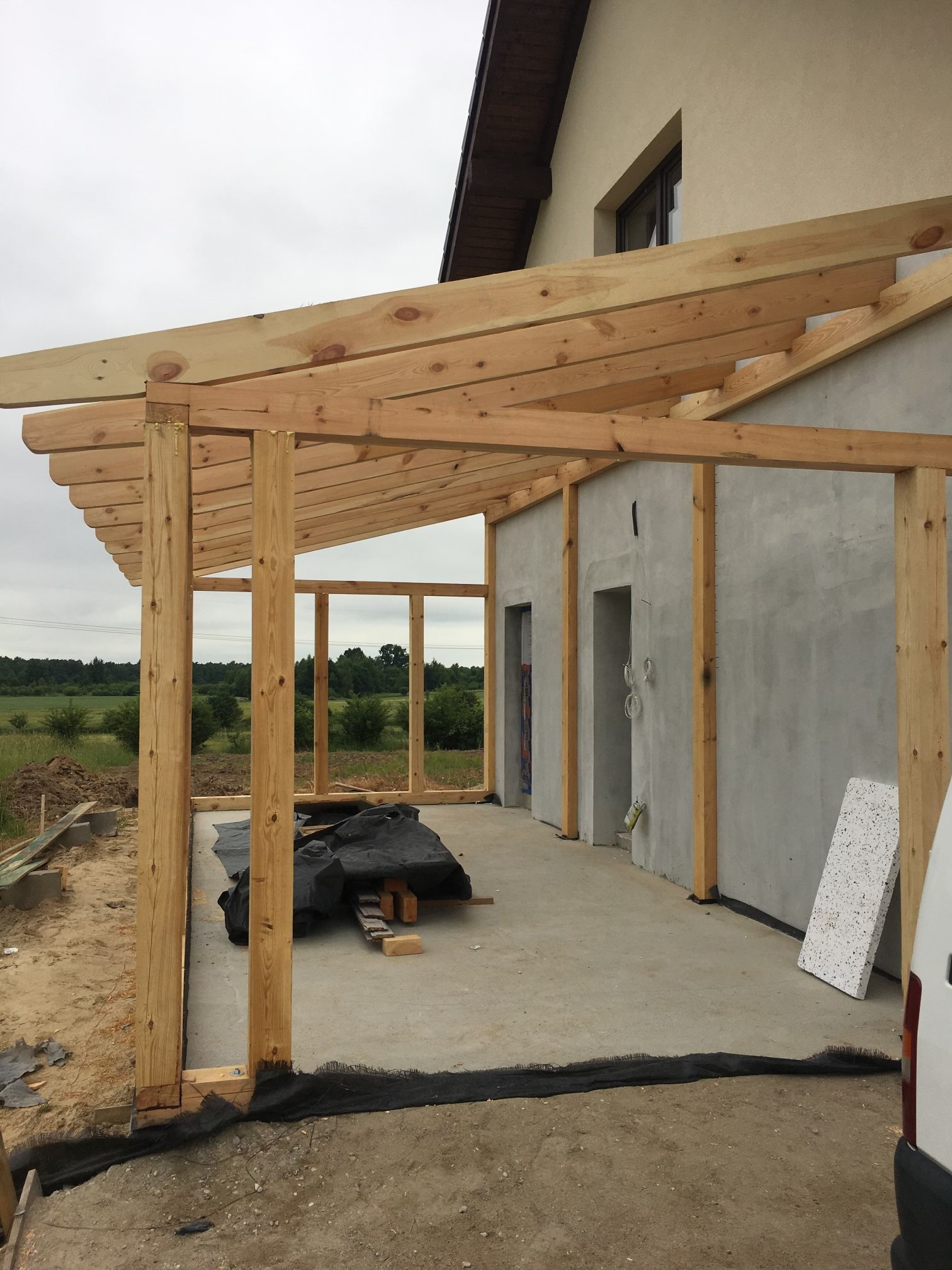 Drewniana konstrukcja w trakcie budowy, prawdopodobnie zadaszenie tarasu przy domu. Widoczne surowe drewno, betonowa podstawa i częściowo otynkowana ściana budynku.