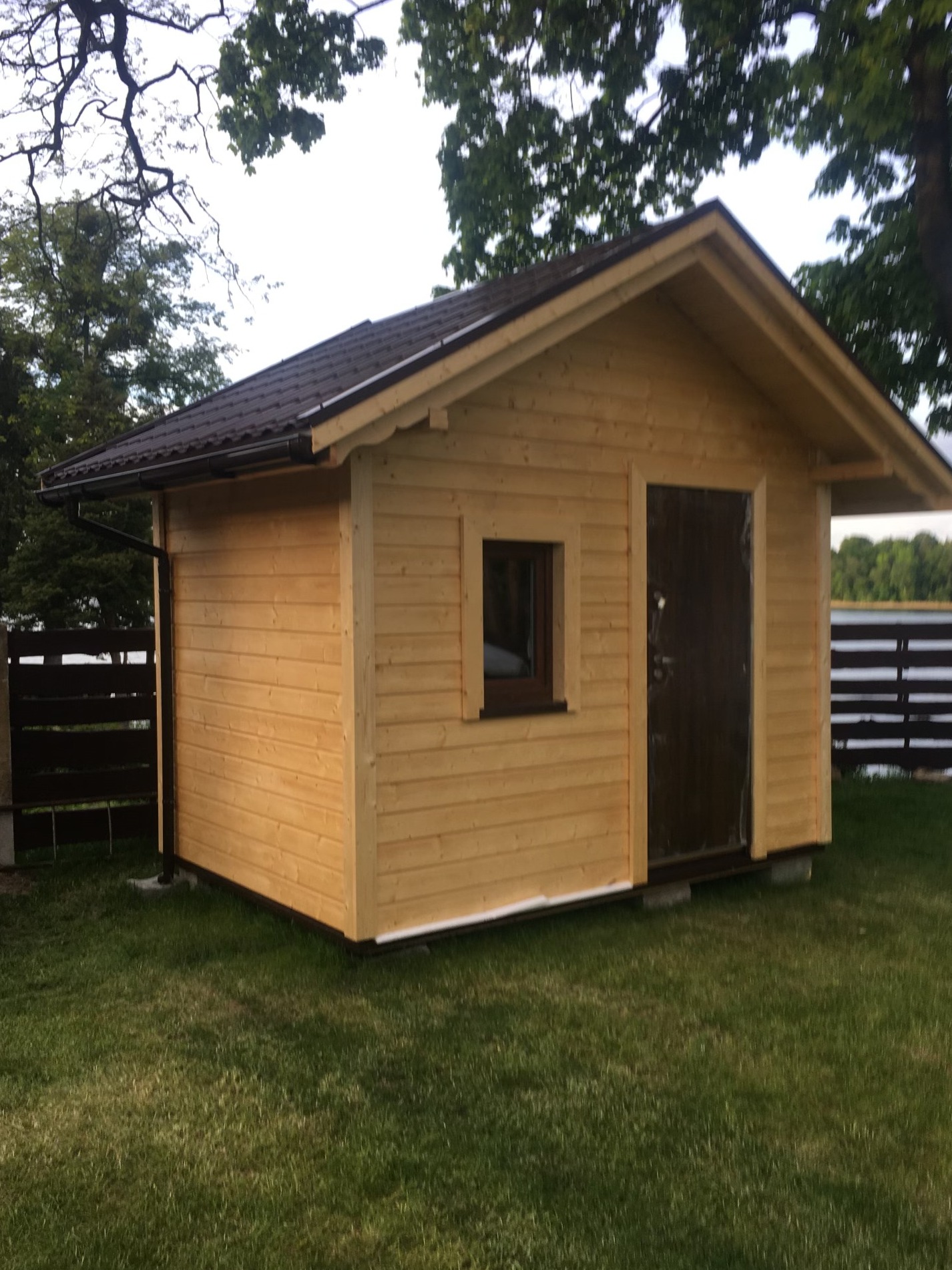 Drewniana altana z brązowym dachem na zielonej trawie, z widocznym oknem i drzwiami. Tło stanowią drzewa i ogrodzenie, tworząc spokojny krajobraz.