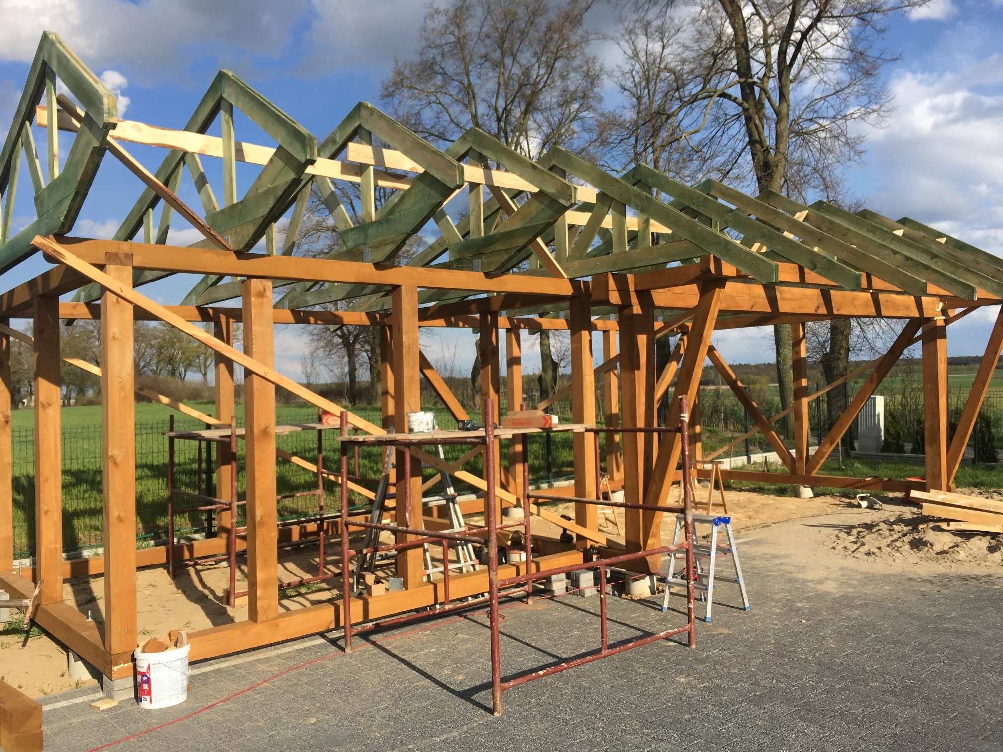 Konstrukcja altany z drewna w trakcie budowy, widoczne rusztowania i elementy więźby dachowej pomalowane na zielono, na zewnątrz, w tle drzewa i niebo.