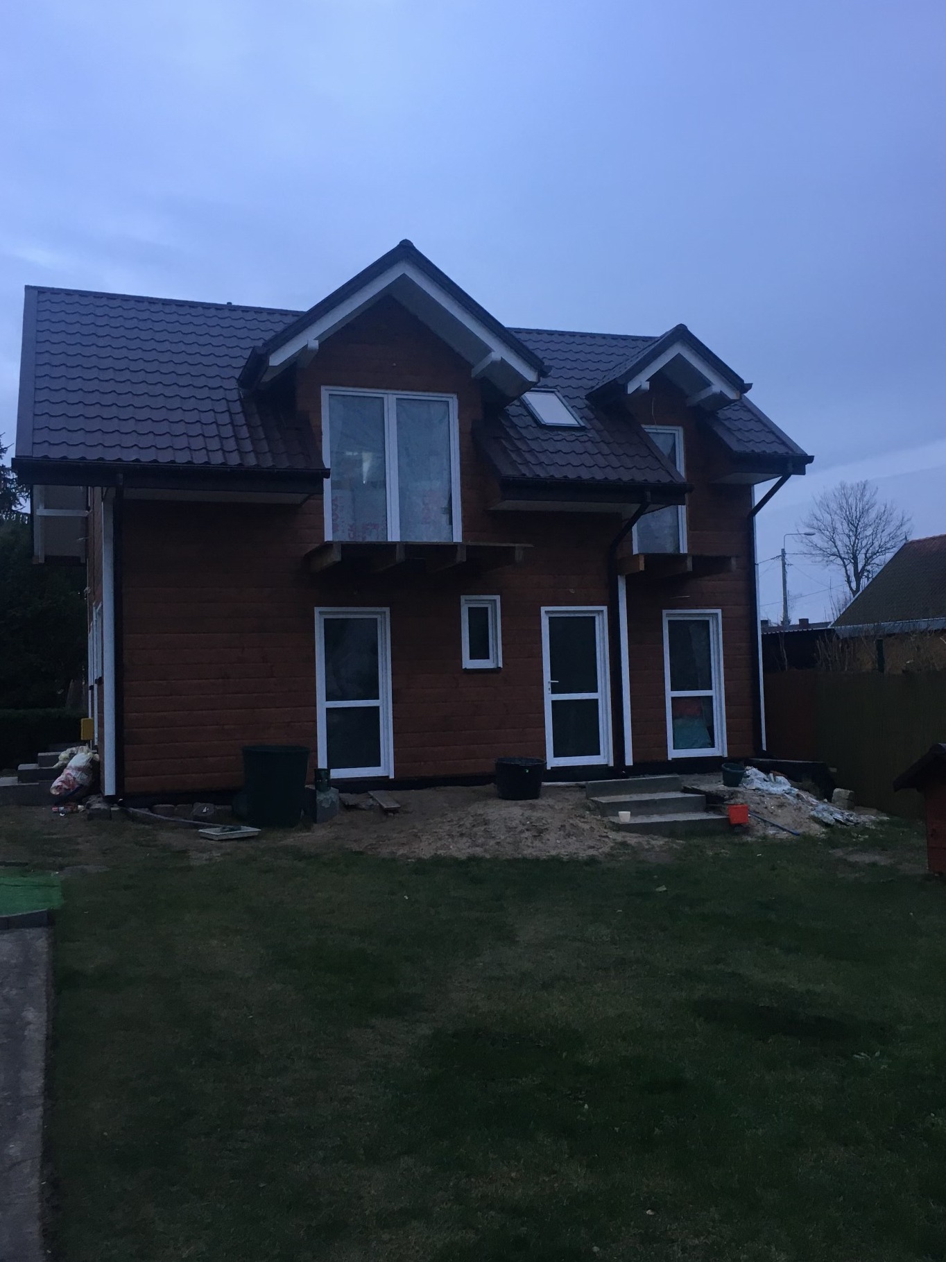 Brunatny dom z brązowym dachem, białe ramy okien i drzwi, otoczony trawnikiem i elementami budowlanymi. Widok z perspektywy ogrodu, pochmurne niebo.