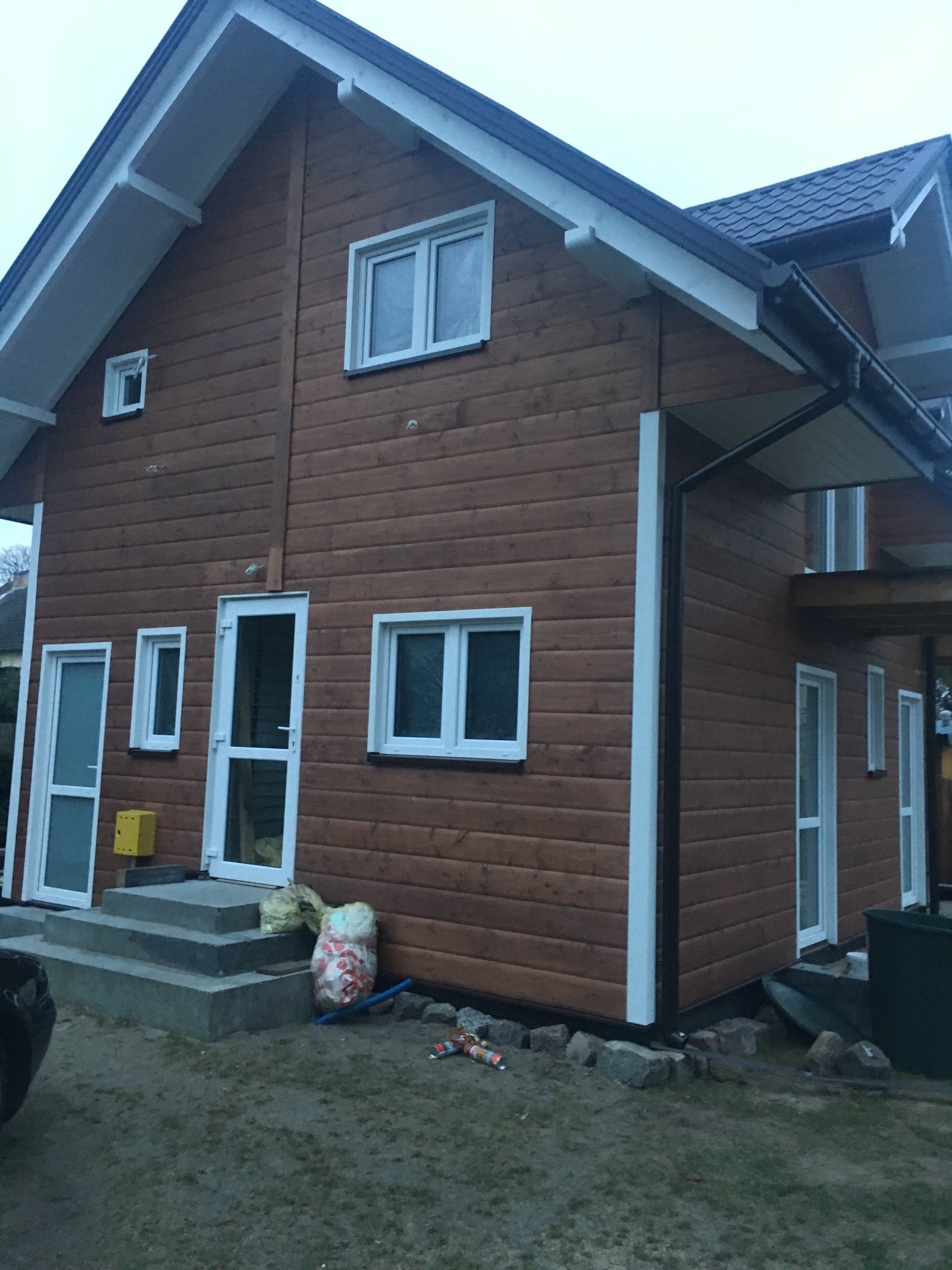 Brązowy dom z drewnianą elewacją i białymi ramami okiennymi, z widocznymi elementami wykończeniowymi. Ujęcie z zewnątrz, z niewielkim fragmentem samochodu po lewej stronie.