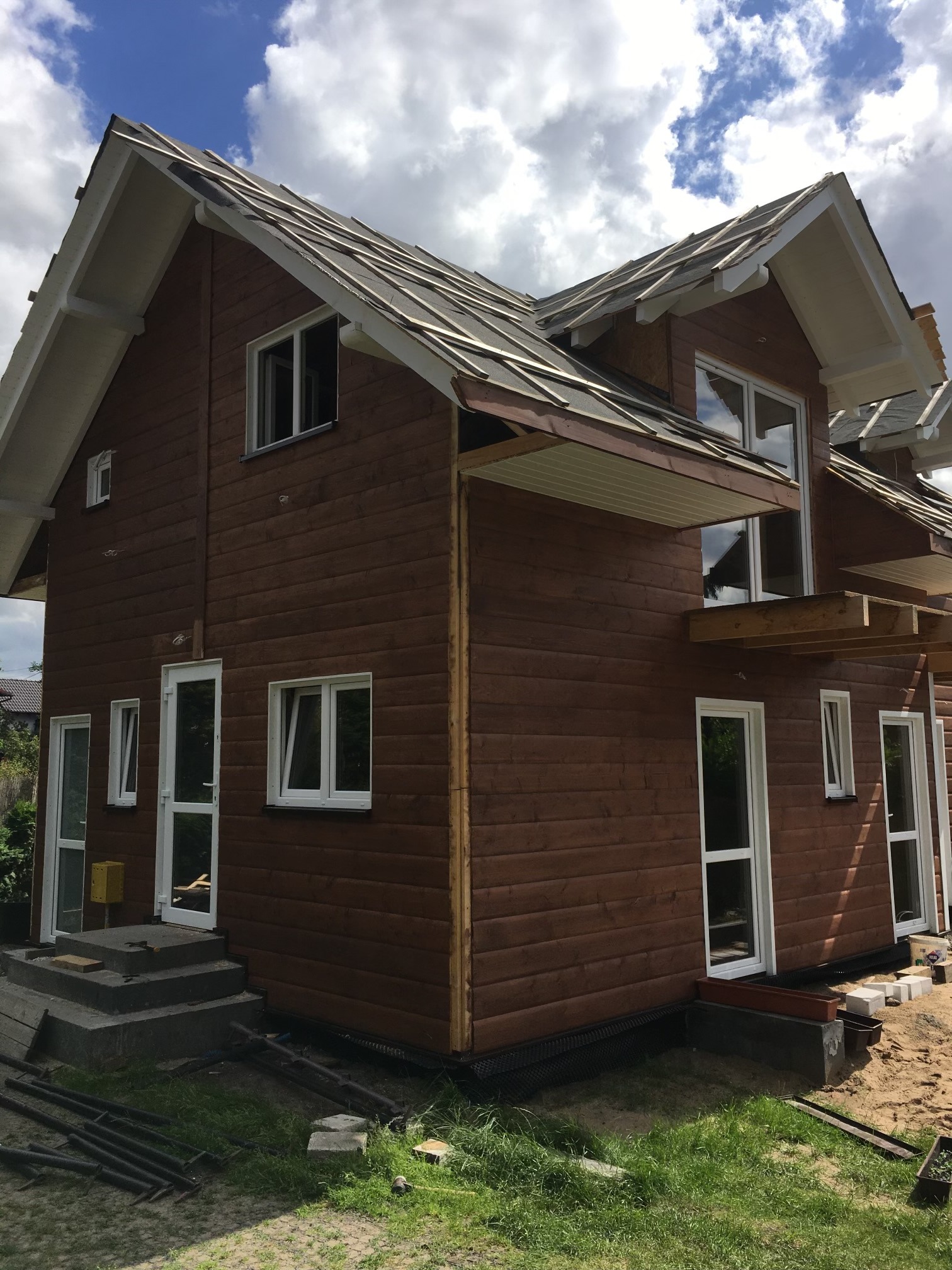 Brunatny dom w trakcie budowy z widocznym ociepleniem dachu i elementami wykończeniowymi. Widoczne schody wejściowe i częściowo zagospodarowany teren wokół.