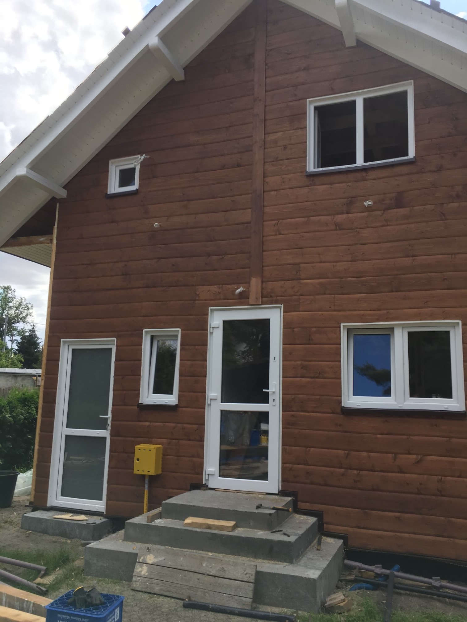 Brunatna elewacja domu z bali, białe ramy okien i drzwi, betonowe schody wejściowe w trakcie budowy, widoczny fragment instalacji gazowej i narzędzi.