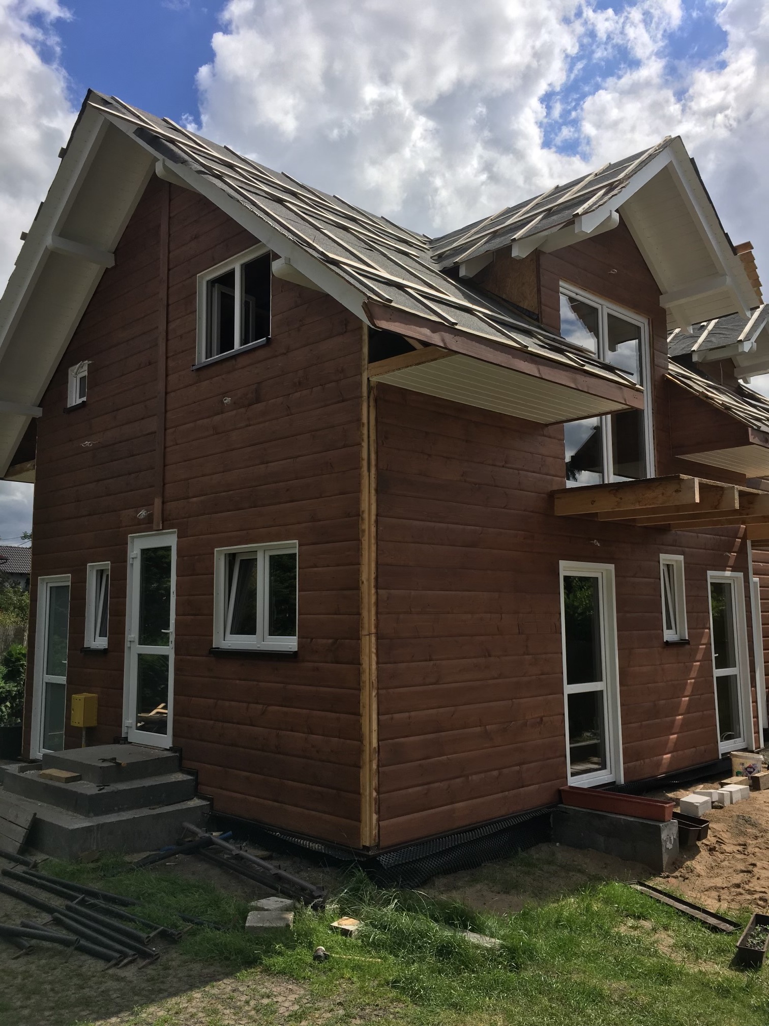 Brązowy dom szkieletowy w trakcie budowy. Widoczne drewniane deski elewacyjne, białe okna i częściowo położony dach. Wokół domu teren budowy.