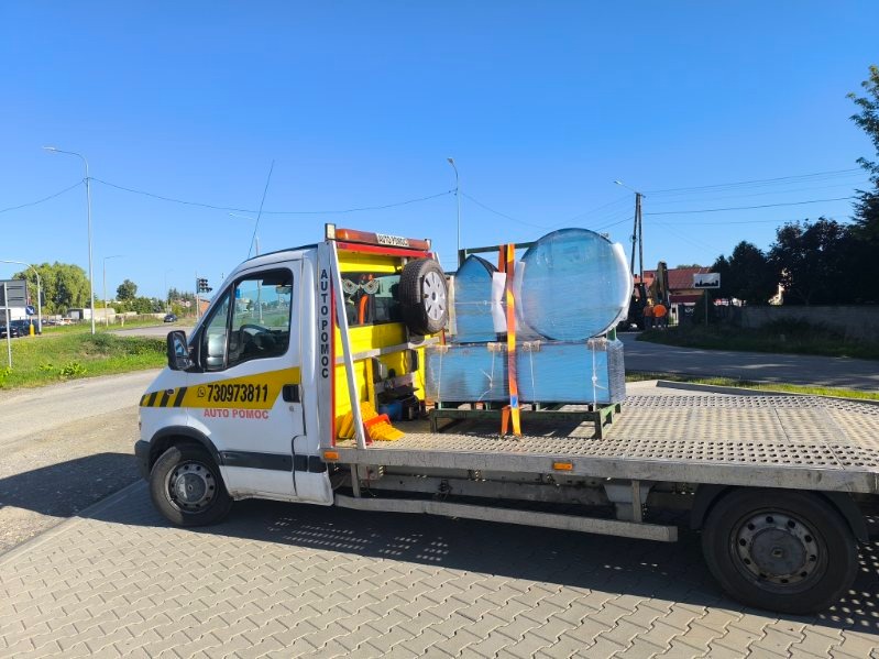 Laweta z platformą przewozi zabezpieczone elementy szklane o różnych kształtach, w tym okrągłe i prostokątne, na tle ulicy i zieleni.