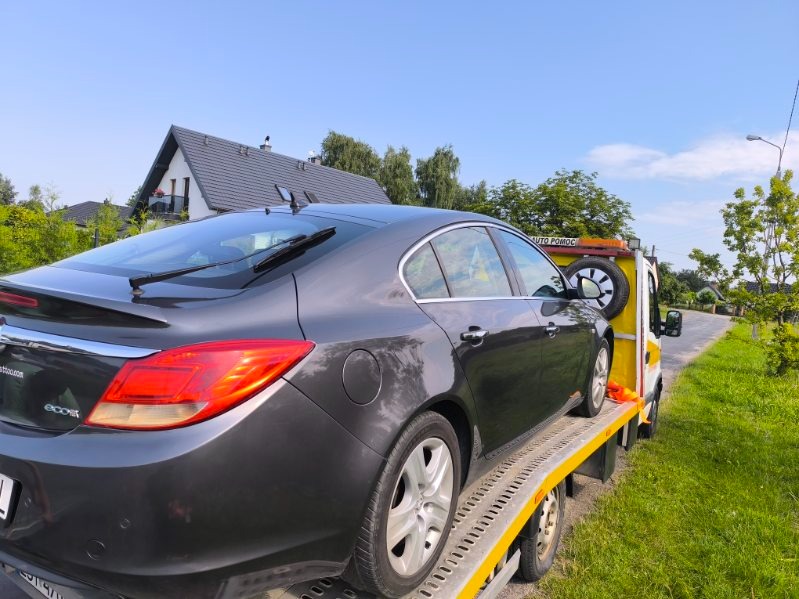 Szary Opel Insignia na żółtej lawecie w słoneczny dzień. Widoczny dom w tle. Auto zabezpieczone pasami.