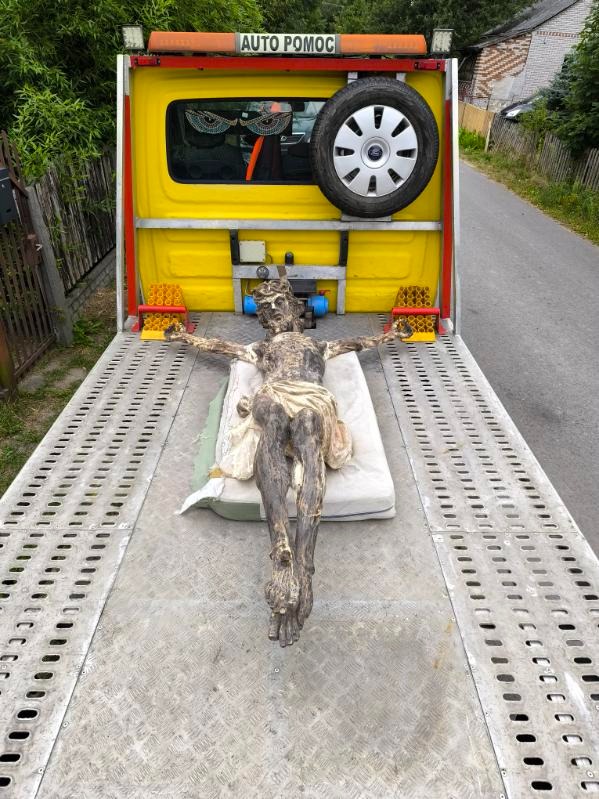 Laweta przewozi rzeźbę Jezusa na krzyżu, umieszczoną na materacu. Żółty samochód pomocy drogowej z zapasowym kołem i napisem 'AUTO POMOC'.