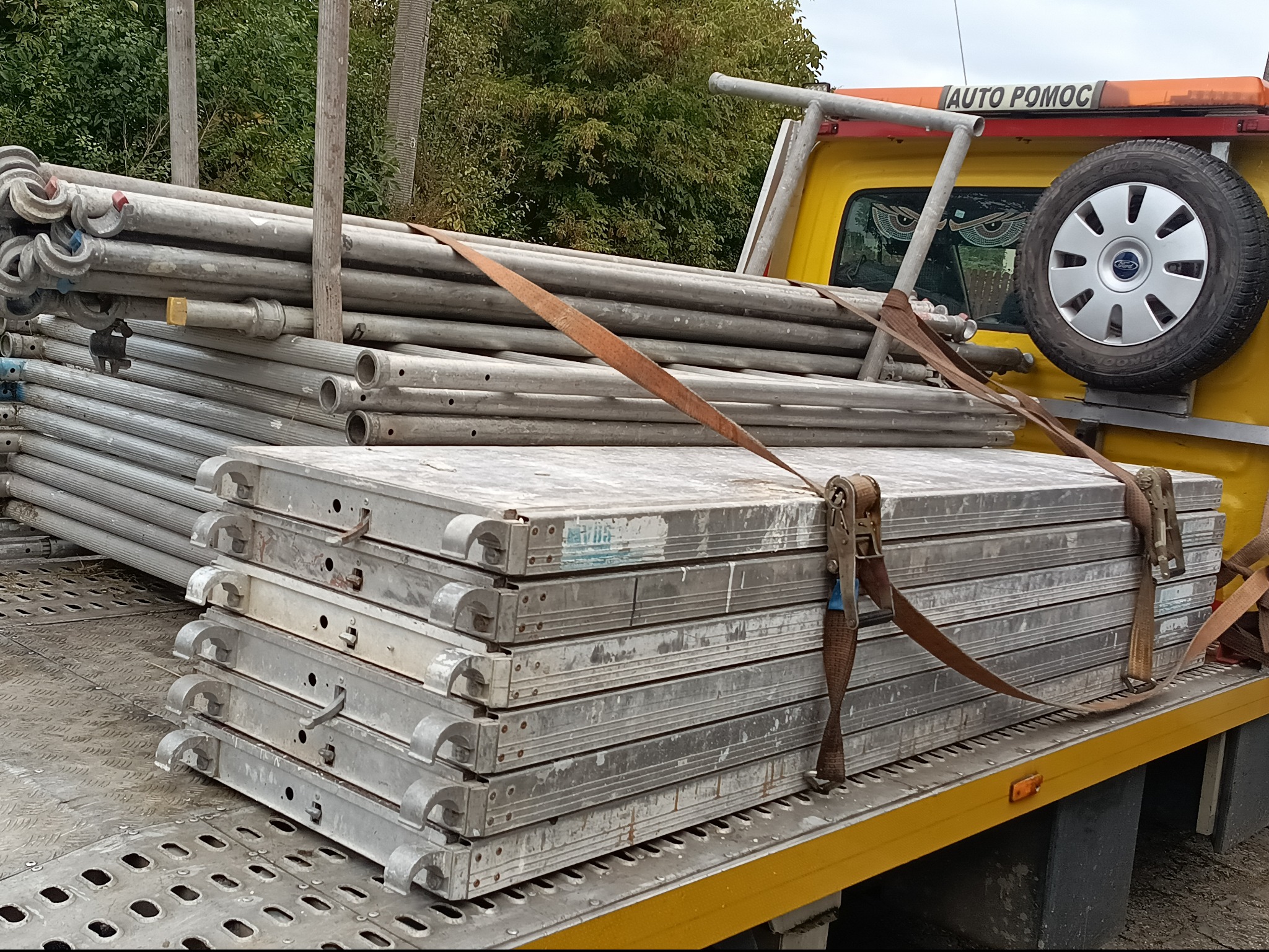 Żółta laweta z napisem 'AUTO POMOC' przewozi zabezpieczone pasami rusztowania budowlane. Widoczne koło zapasowe i elementy karoserii pojazdu.