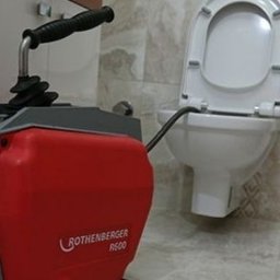 PERFECT CLEAN DANIEL MICHOŃ - Toaleta z otwartą klapą i urządzenie Rothenberger R600 do udrażniania rur, z widoczną giętką rurą wewnątrz muszli, na tle beżowych płytek.