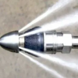 Dysza udrażniająca rury pod ciśnieniem, woda tryska z otworów, metalowa konstrukcja, szary beton w tle. Widok z bliska.